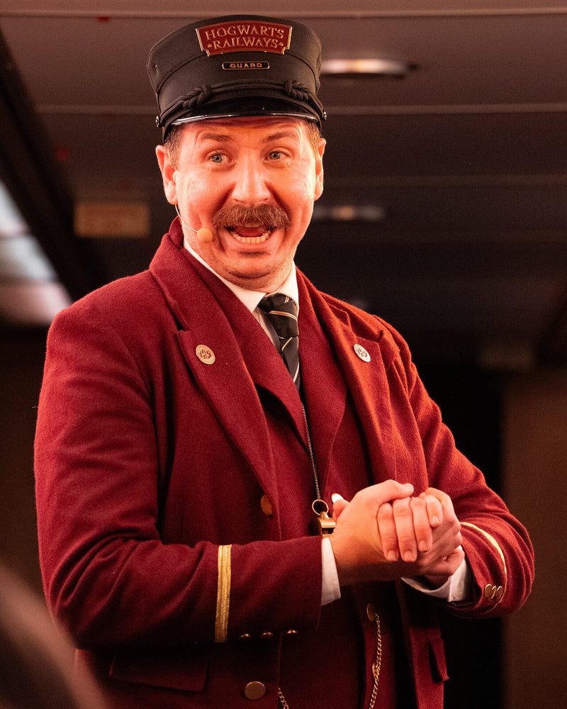 A conductor in a red wool uniform and braided cap aboard the Hogwarts Express Adventure
